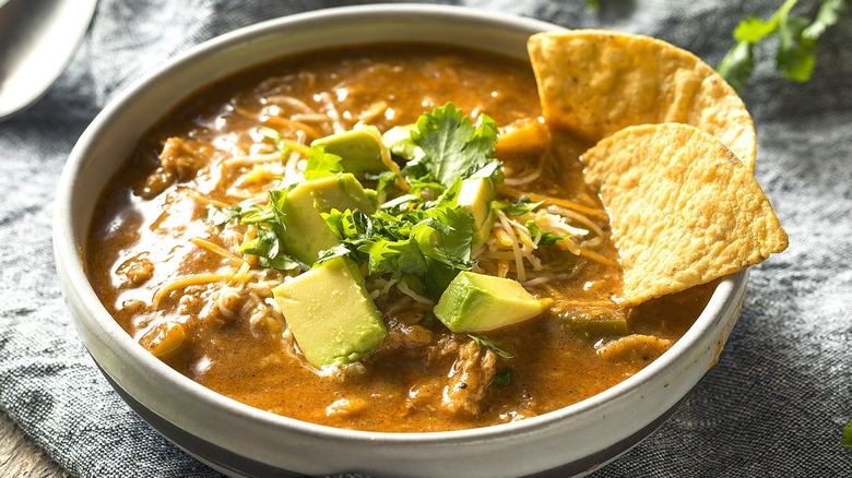 A bowl of creamy chicken soup with beans, fresh cubed avocado, and tortilla chips over a grey tablecloth