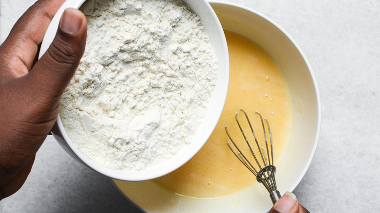 A hand pouring flour into cake batter, the other hand working the whisk to incorporate it.