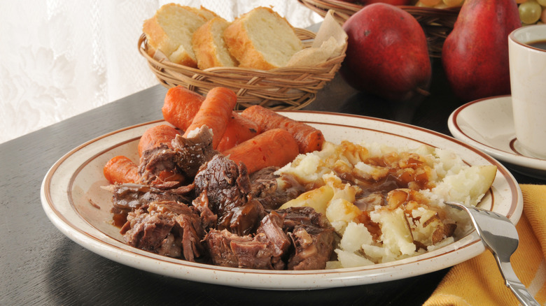 Pot roast with carrots and gravy-covered mashed potatoes next to pears and a wicker basket filled with bread