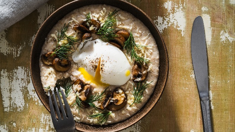 A bowl of savory oatmeal with a poached egg, sautéed mushrooms, Parmesan cheese, and sprigs of dill sits on a rustic table.