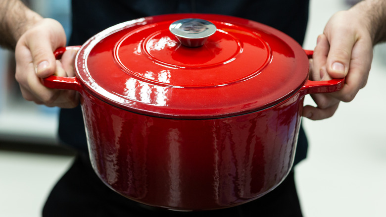 Two hands holding a red, cast iron Dutch oven.