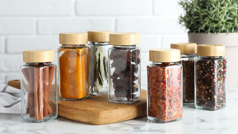 A variety of spices in clear jars with wooden caps sitting on a marble countertop.