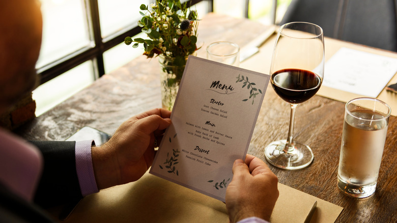 A person perusing a restaurant's menu at a table with a glass of red wine and water on the side.