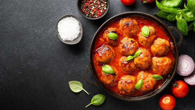A dish of meatballs and red sauce with basil appears with seasonings and veggies in the frame.