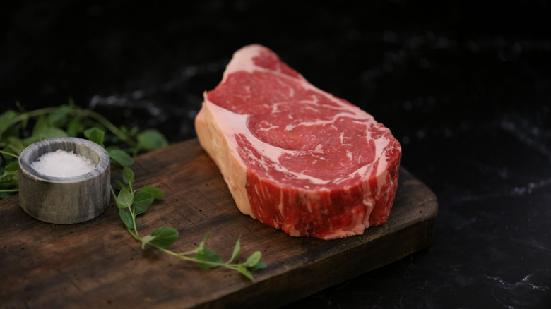 A raw ribeye steak on a cutting board with salt and herbs