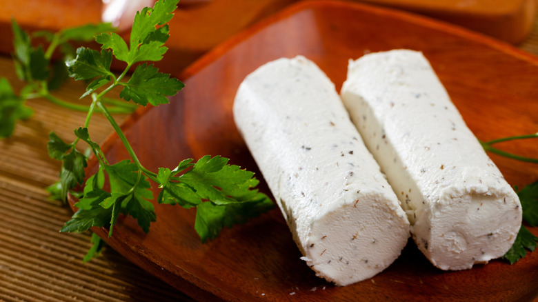 Two logs of fresh goat cheese with pepper and herbs on a wooden tray, a sprig of parsley to the side.