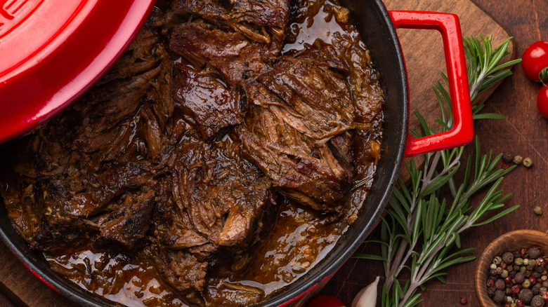 Close up of cooked pot roast in a deep red pan on wood cutting board with sprigs of rosemary