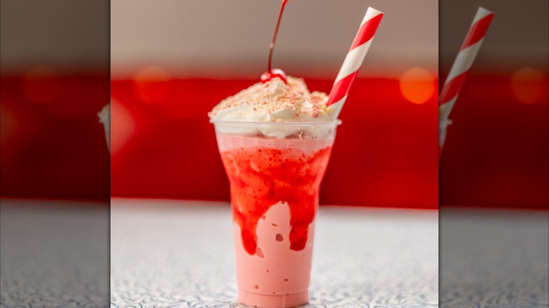 Strawberry milkshake with whipped cream, a cherry, and a crumbly topping in a plastic cup with a red and white straw