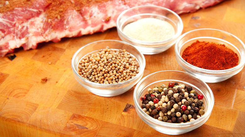 Little bowls of spices for a dry rub are arranged in front of a rack of ribs