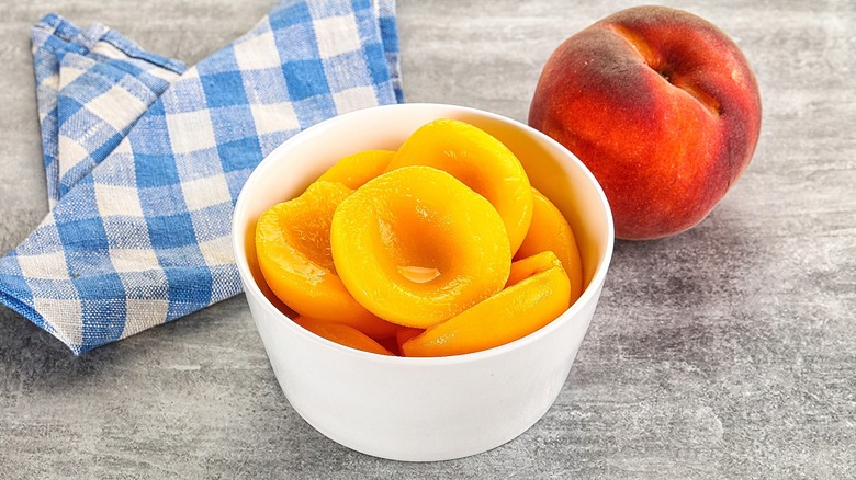 Halved canned peaches in a small white bowl next to a whole peach and a folded blue and white checkered cloth napkin