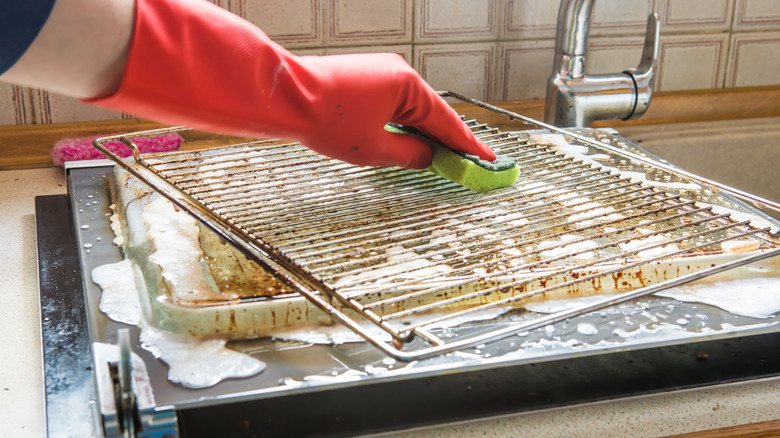 A person's gloved hand is seen scrubbing an oven rack over a kitchen sink.