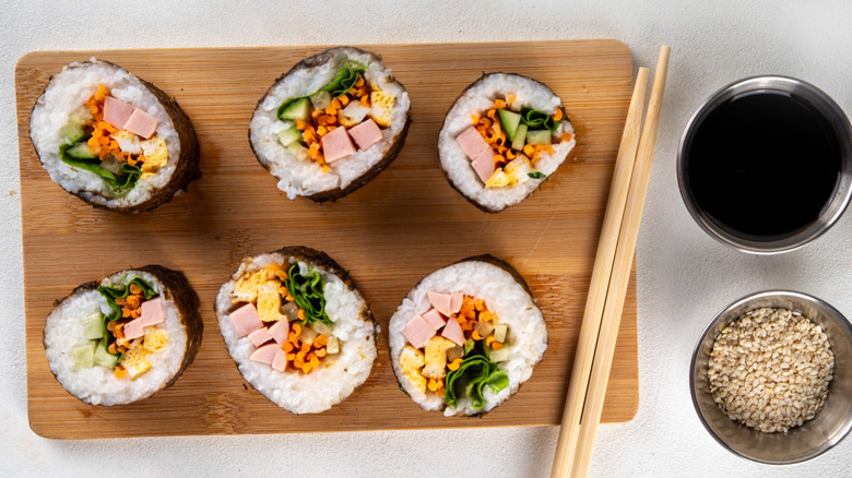 Kimbap on a wooden cutting board with chopsticks next to containers of soy sauce and sesame seeds