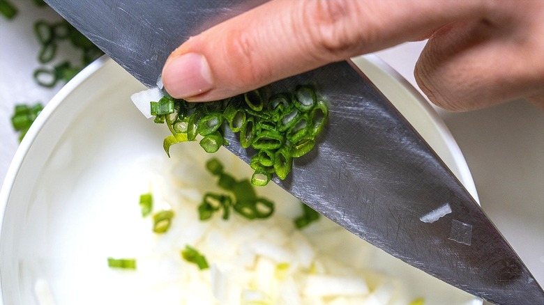 Finger swiping chopped green scallions off the blade of a knife into a bowl