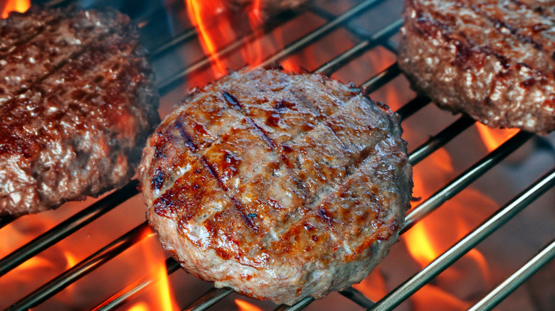 Three perfectly-formed hamburgers on a hot grill, the flames lapping at them from below.