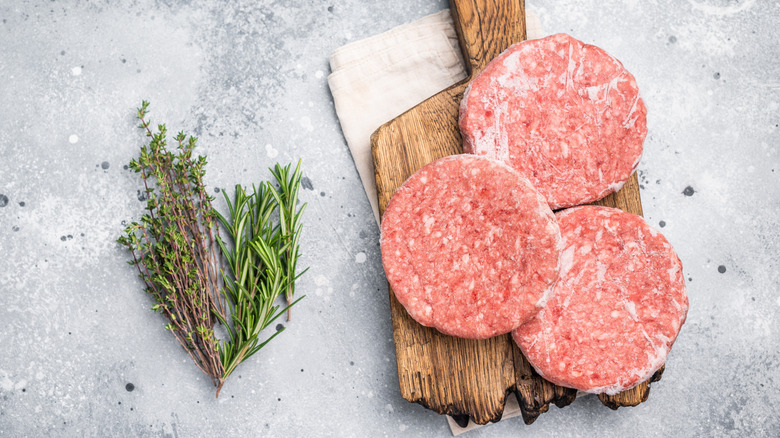 Frozen burger patties on a wooden board next to springs of herbs