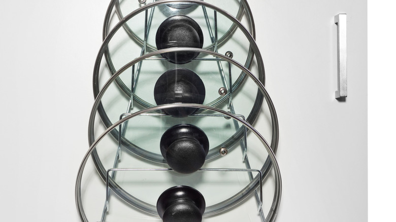 A hanger with glass lids for cooking pots hung on the door of a kitchen cupboard.