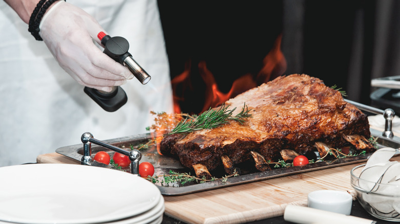 A chef blowtorching a prime rib roast on a serving tray.