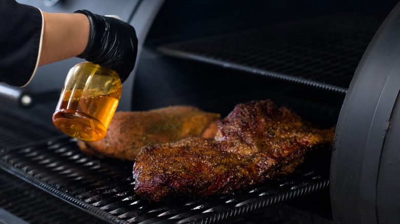 A hand spraying liquid from a spray bottle over meat on a grill.