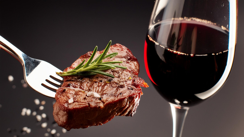 Bite of steak with rosemary on a fork next to a glass of red wine