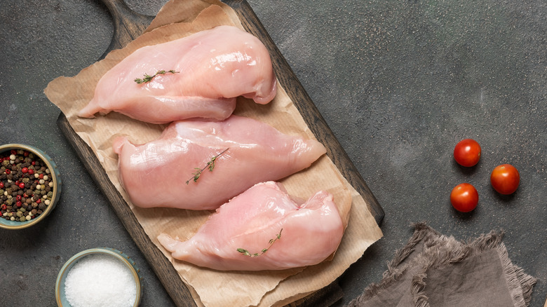 Three pieces of raw chicken breast placed atop a parchment paper on a wooden cutting board.