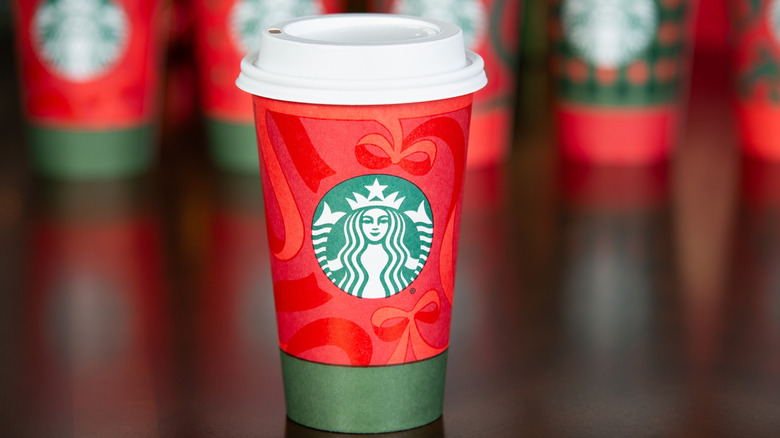 2025 Starbucks Christmas cup in focus with another cup and a lit Christmas tree with presents blurred in the background