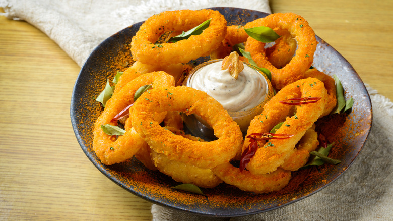 A homemade bowl of onion rings on a wooden countertop, with a dairy dipping sauce, topped with herbs and spices.
