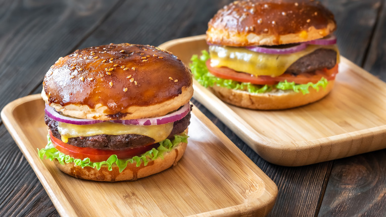 Two glazed cheeseburgers on wooden plates in a sesame bun, topped with red onions, lettuce and tomato on the bottom.
