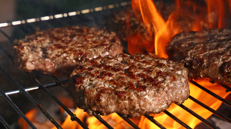 A close-up of three hambugers with beautiful browning on a hot grill, a flame in the background.