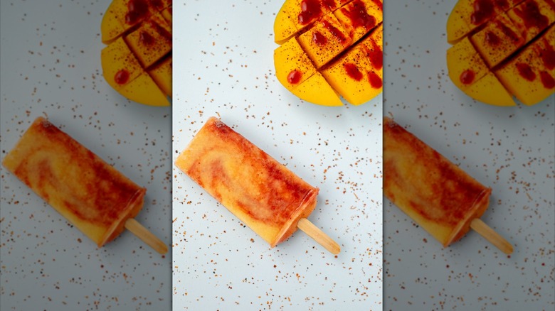 Mango and Tajín popsicle next to a mango fruit covered with Tajín and Chamoy drizzle