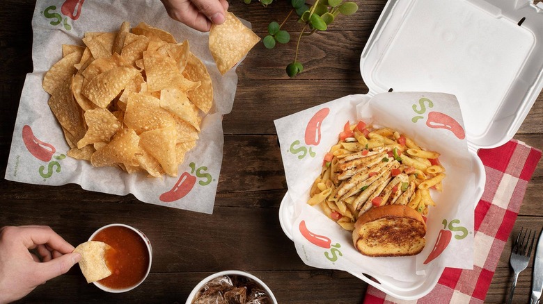 Overhead shot of a Chili's to-go box with burgers and fries, a basket of chips, and people's hands dipping chips into salsa