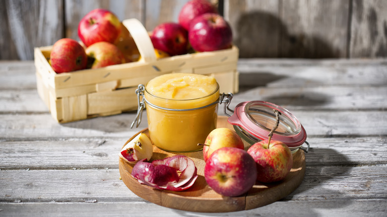 A jar of appleasuce appears on a wooden tray with more apples in a rustic setting.