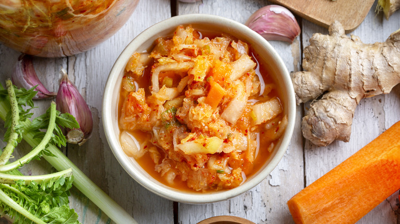 Bowl of homemade kimchi surrounded by garlic cloves, a peeled carrot, whole ginger, and jar of kimchi.