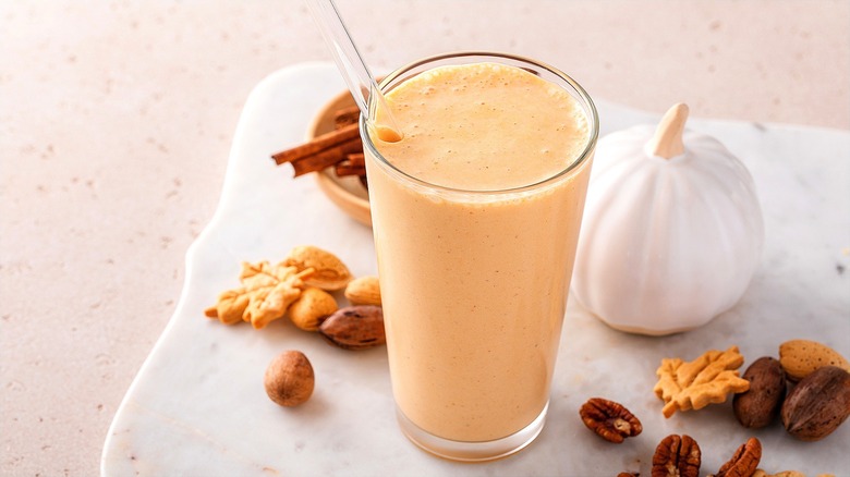 Pumpkin smoothie in a tall glass with a clear straw surrounded by nuts and small leaf-shaped cookies