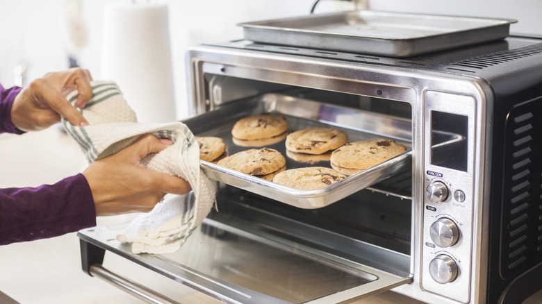 A cook removing a tray of chocolate chip cookies from a toaster oven