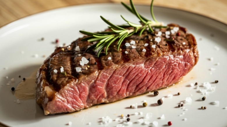 A perfectly seared flank steak garnished with rosemary, peppercorns, and salt sitting on a white plate ready to be served