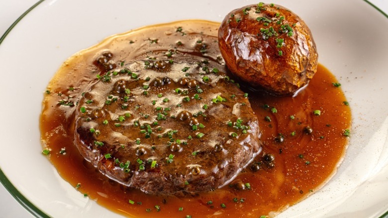 A round cut of steak smothered in gravy with herbs next to a golden baked potato