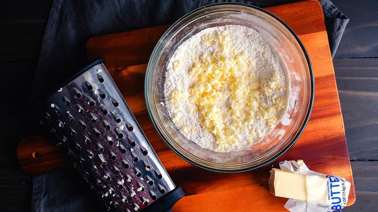 Grated butter over flour in a clear mixing bowl on a wooden cutting board