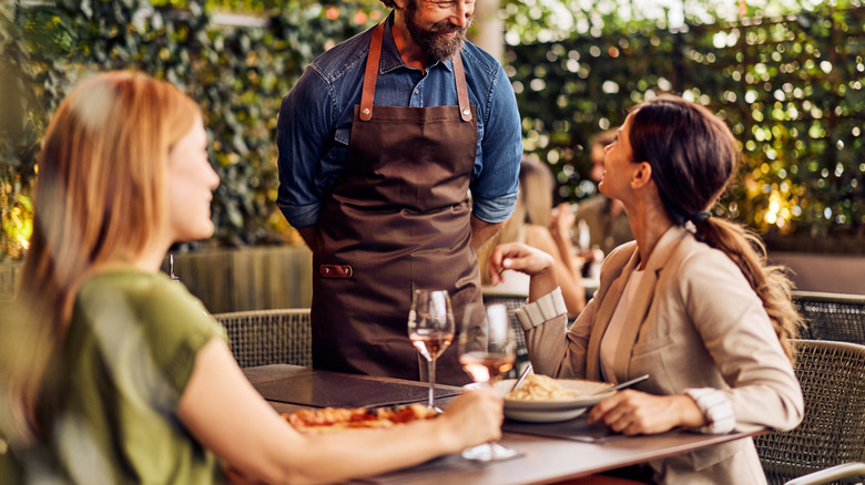 Two women sitting at a table while a server talks to one of them with a smile.