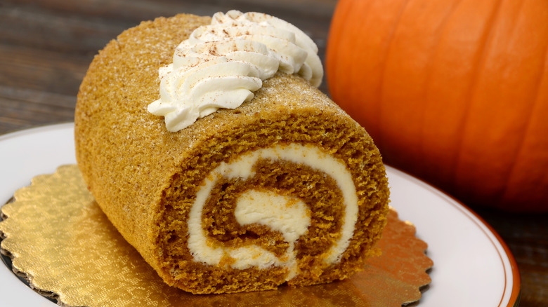 Pumpkin roll on a golden platter with whipped cream topping next to a pumpkin