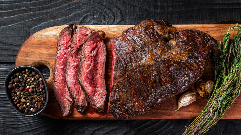 A sliced steak on a cutting board that appears to be cooked medium rare, with peppercorns, garlic, and herbs on the side