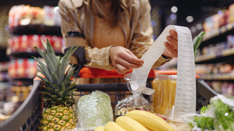 A person reads a printed grocery store receipt over a cart filled with food.