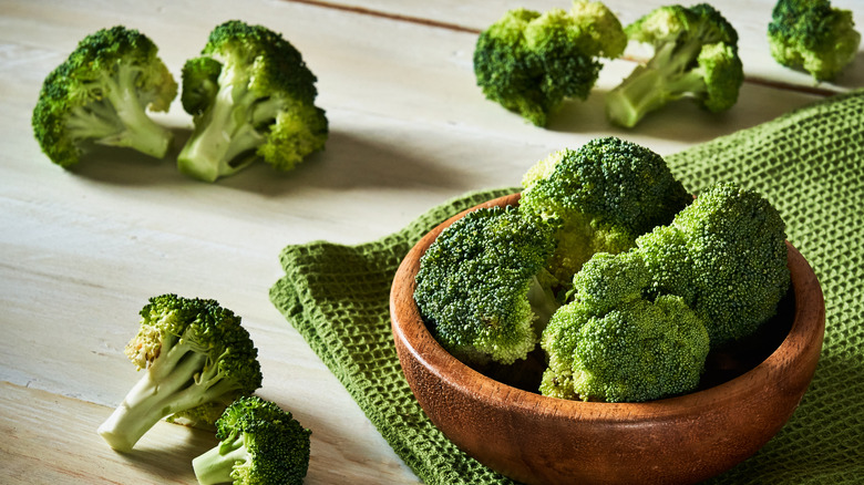 Fresh broccoli florets on a white, wooden table, some in a bowl over a green tea towel.
