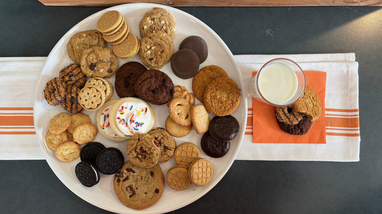 White plate of assorted Aldi cookies on orange and white kitchen towel next to milk and cookies