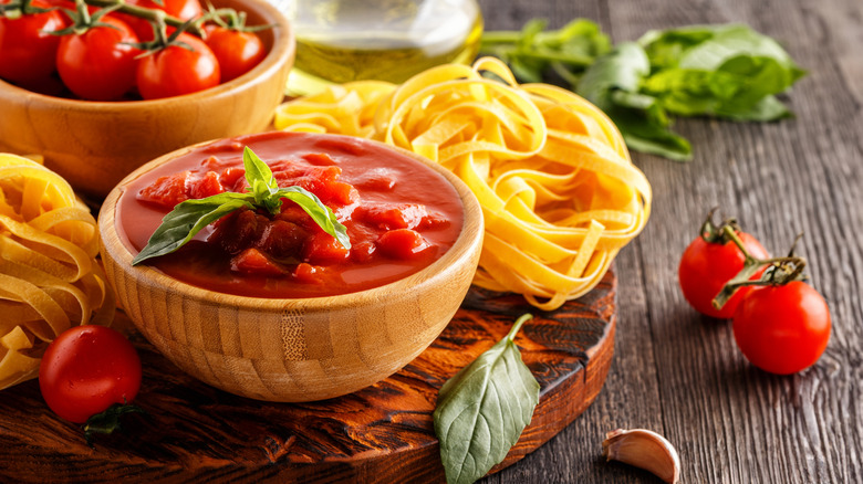 A wooden bowl of tomato sauce is pictured near nests of noodles and a bowl of tomatoes on the vine.