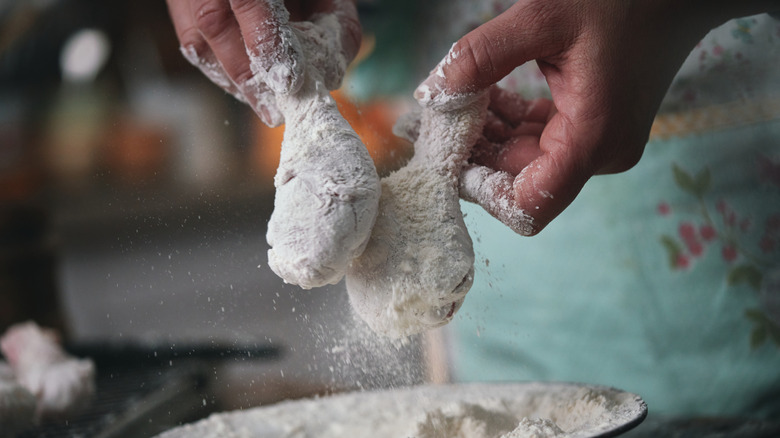 Hands dipping chicken drumsticks in flour.