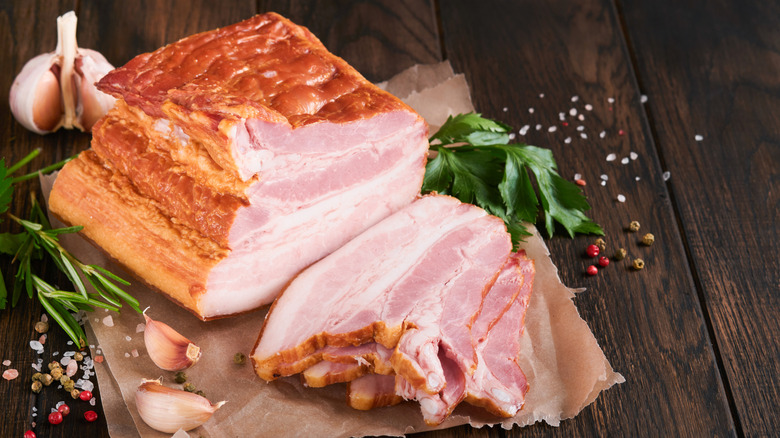 A whole smoked slab of bacon on butcher paper, some strips sliced off, surrounded by garlic cloves, salt, and peppercorns.