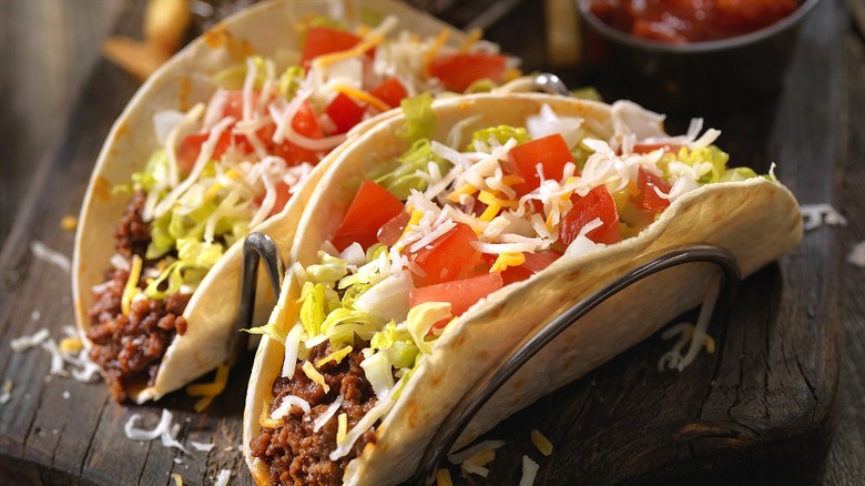 Two soft-shell tacos filled with ground beef, shredded cheese, lettuce, and chopped tomatoes in a wire stand