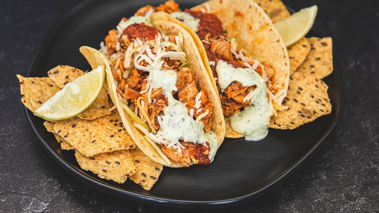 Two chicken tacos with shredded cheese and cream sauce on a black plate with whole grain chips and wedges of lime