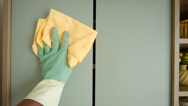 Gloved hand cleaning kitchen cabinet with a yellow cloth