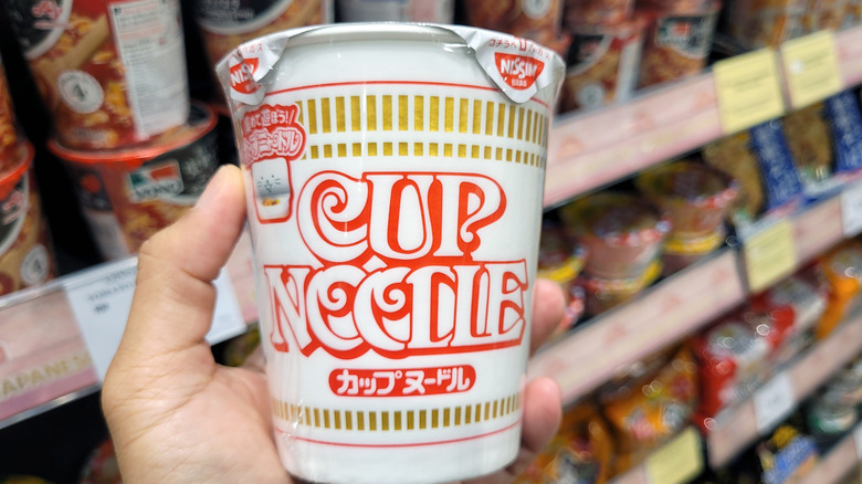 A hand holding a Nissin Cup Noodle at a supermarket, along the instant ramen aisle.